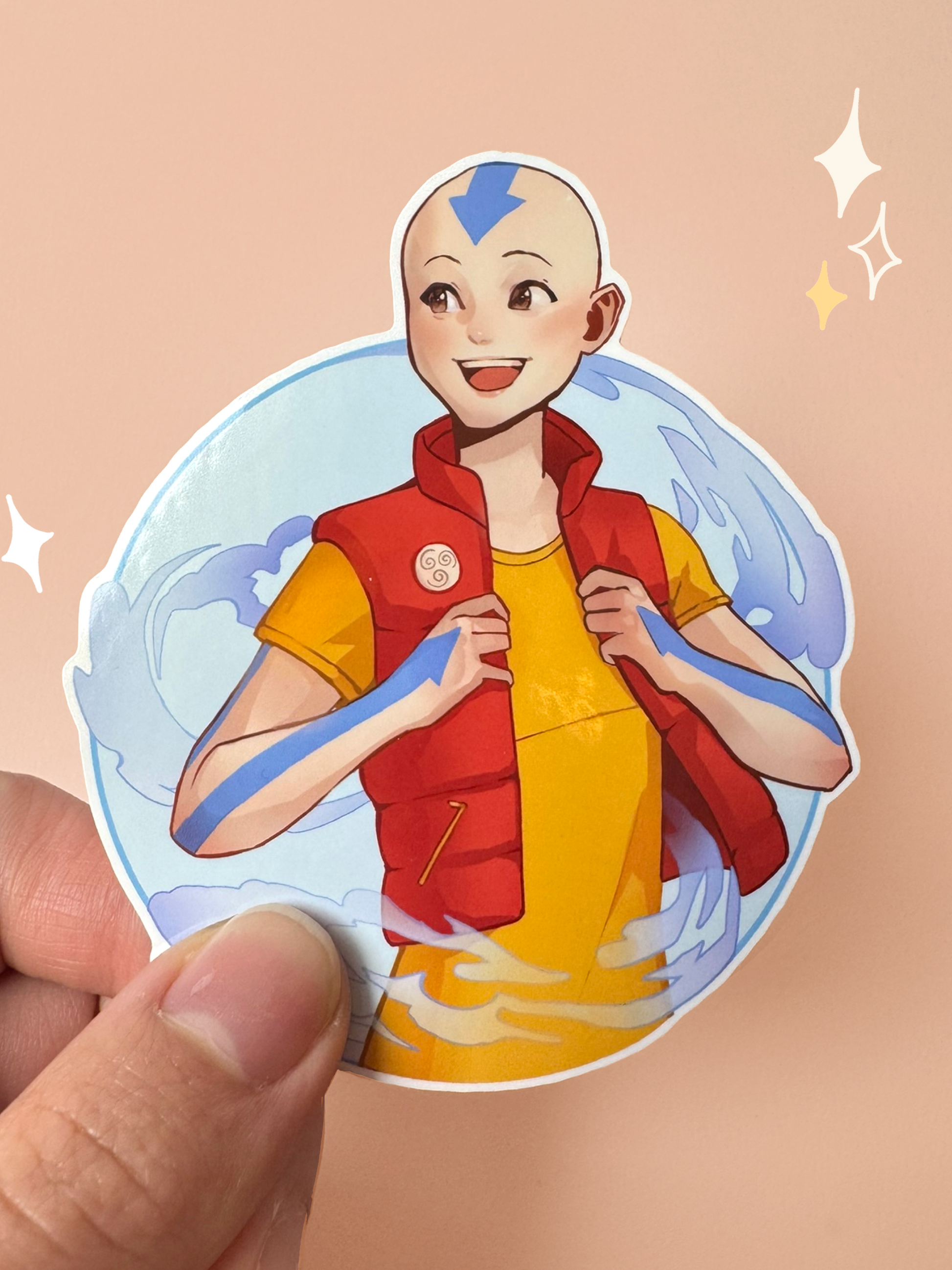 Die-cut vinyl sticker of Aang from Avatar: The Last Airbender in a modern outfit — yellow t-shirt with red puffer vest displaying the Air Nomad insignia, blue air-arrow tattoos visible on his head and arms, with airbending swirls around him in a circular frame, held between fingers against a peach background.
