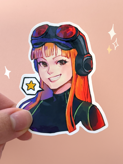 Die-cut vinyl sticker of Futaba Sakura (Oracle) from Persona 5 Royal, grinning with red goggles and headphones over long orange hair and a star speech bubble, held between fingers against a peach background.