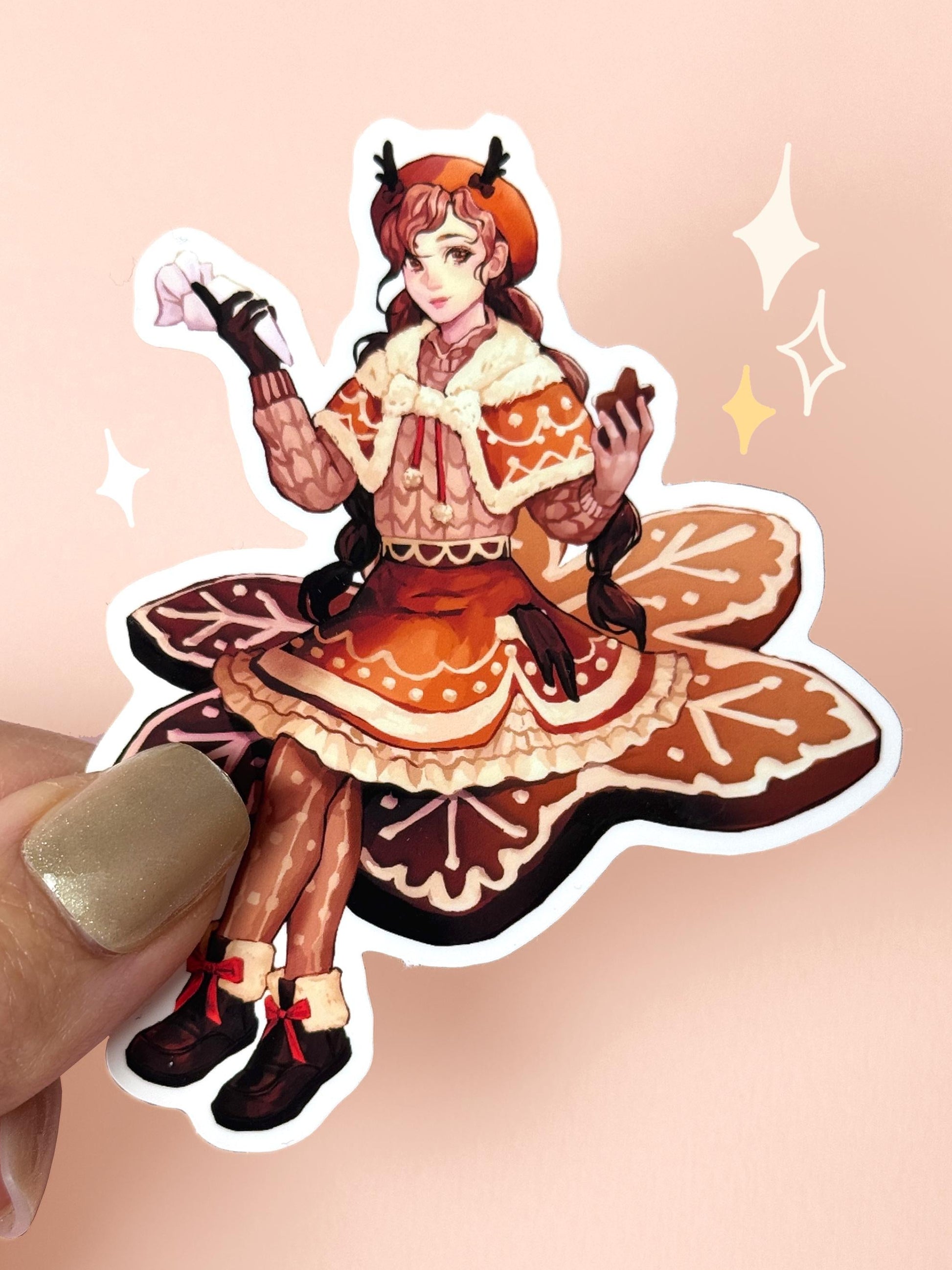 Die-cut vinyl sticker of a girl in a gingerbread-themed outfit — brown dress and cape iced with white frosting trim, antler beret, sitting on a giant gingerbread cookie, holding a smaller cookie, held between fingers against a peach background.