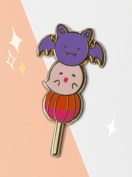 Hard enamel pin shaped like a dango skewer — a winged purple bat on top, a pale ghost in the middle, and an orange-and-pink pumpkin at the base, all on a gold stick.