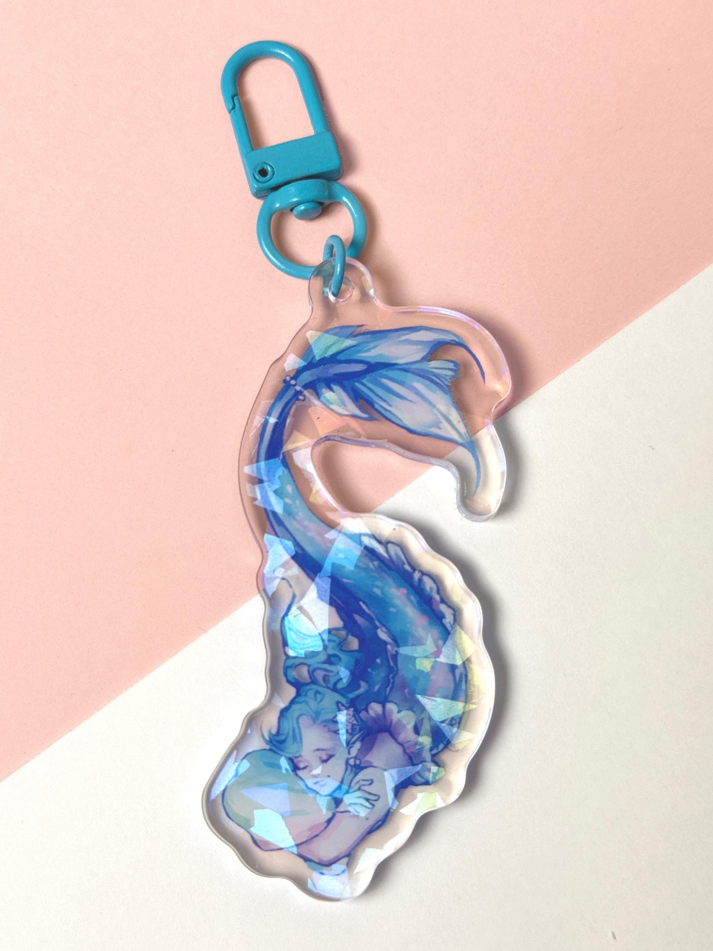 Holographic acrylic keychain of a sleeping mermaid with flowing blue hair and a shimmering blue tail curled around her, with a broken-glass iridescent finish and a blue clasp.