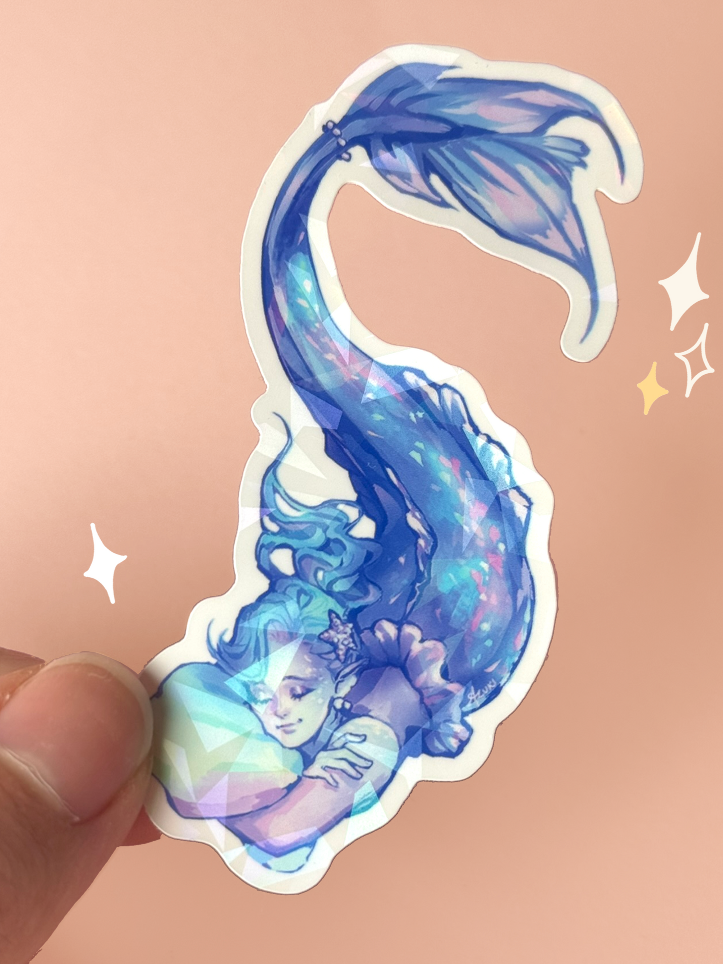 Die-cut holographic vinyl sticker of a dreamy mermaid with flowing blue hair and a shimmering blue-and-pink tail, sleeping while cuddling a seashell pillow, with a broken-glass holographic finish, held between fingers against a peach background.