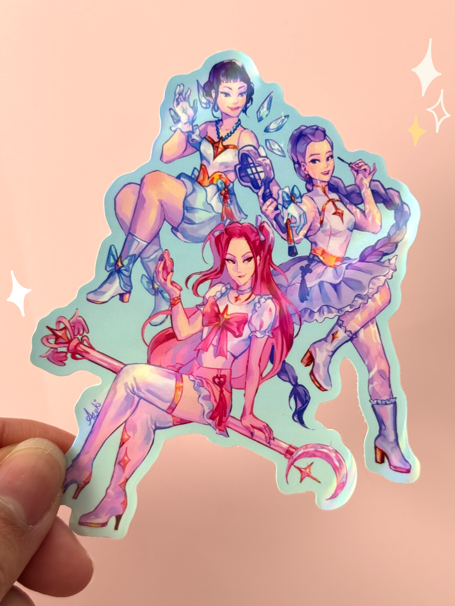 Die-cut holographic vinyl sticker of the three members of HUNTR/X from K-Pop Demon Hunters reimagined as magical girls — Rumi with pink hair holding a crescent moon staff, Mira with long purple hair holding a mascara wand, and Zoey with short black hair holding a tennis-racket weapon — all in pastel ruffled magical girl outfits, held between fingers against a peach background.
