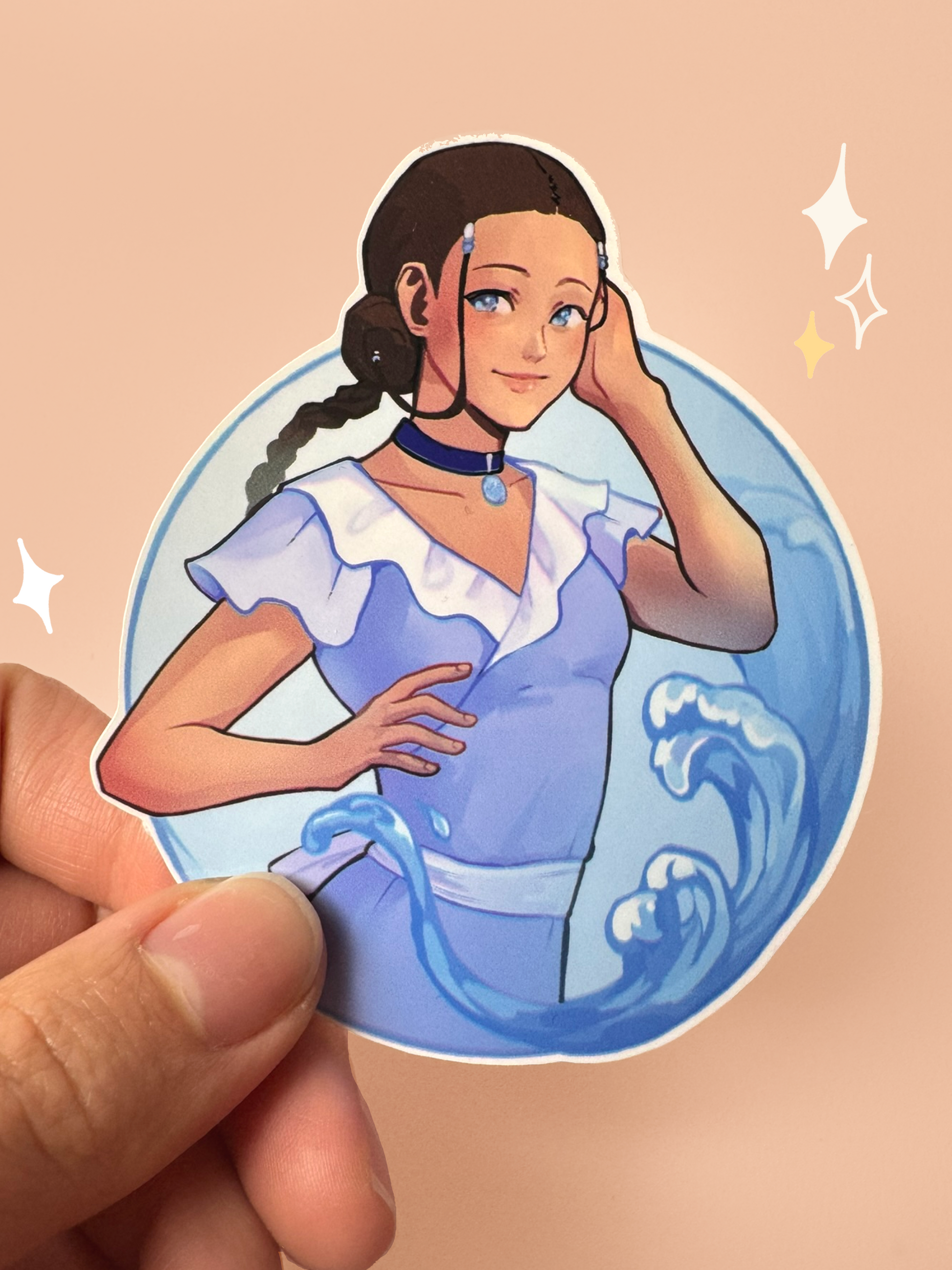 Die-cut vinyl sticker of Katara from Avatar: The Last Airbender in a modern outfit — purple ruffled sundress with a white belt, her signature hair loopies and side braid, a blue Water Tribe choker pendant, with waterbending splashes around her in a circular frame, held between fingers against a peach background.