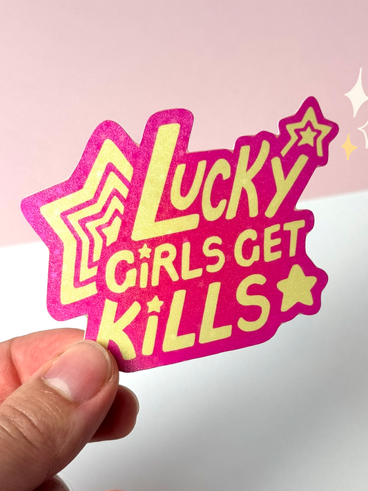Die-cut holographic vinyl sticker with the text "Lucky Girls Get Kills" in yellow bubble letters with stars on a glittery pink background, held between fingers against a pale background.