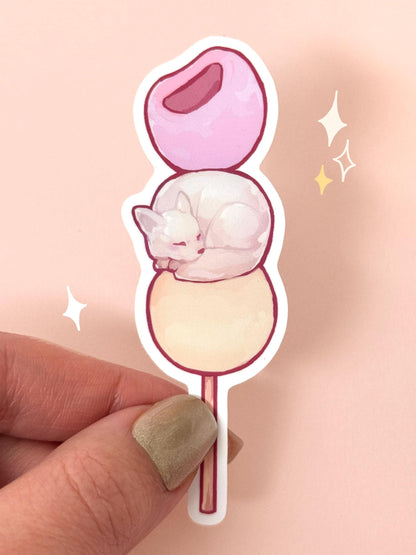 Die-cut soft-touch vinyl sticker of a sleepy white American Eskimo dog curled up on the middle ball of a three-mochi dango skewer in pastel pink, white, and cream, held between fingers against a peach background.