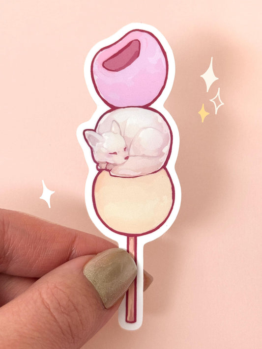 Die-cut soft-touch vinyl sticker of a sleepy white American Eskimo dog curled up on the middle ball of a three-mochi dango skewer in pastel pink, white, and cream, held between fingers against a peach background.