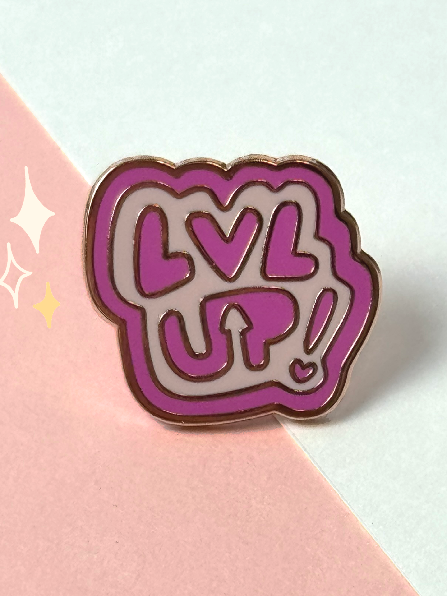 Pink hard enamel pin with bubbly handwritten "LVL UP!" lettering outlined in gold, with a little heart, on a pink and mint background.