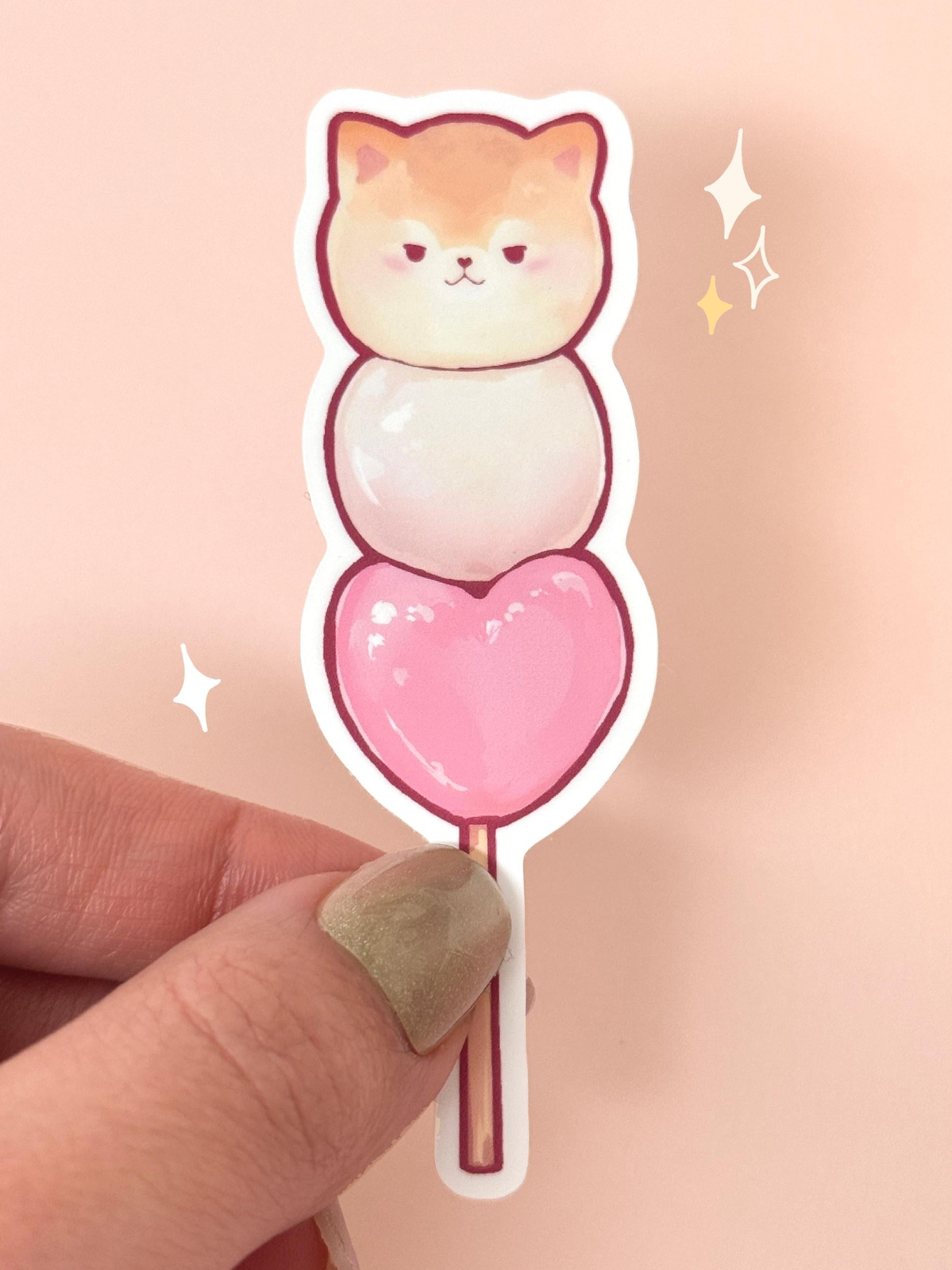 Die-cut soft-touch vinyl sticker of a grumpy orange Pomeranian as the top of a dango skewer, with a white mochi ball in the middle and a pink heart-shaped mochi at the bottom on a stick, held between fingers against a peach background.