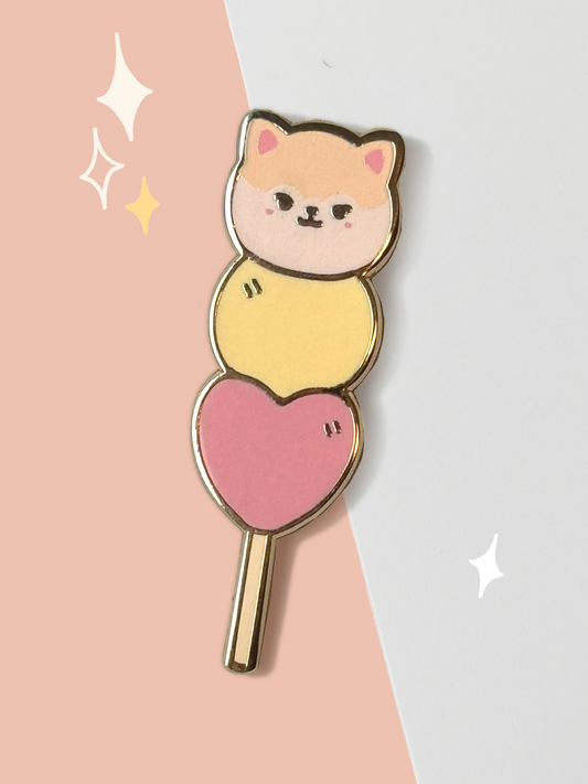 Hard enamel pin shaped like a dango skewer — May the Pomeranian with cream ears and pink cheeks as the top mochi, a yellow mochi in the middle, and a pink heart at the base, all on a gold stick.