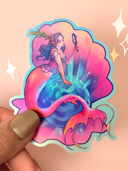 Die-cut rainbow holographic vinyl sticker of a glamorous mermaid with purple hair and a pink-and-orange tail, holding a hand mirror beside a glowing teal pearl in a clam shell, held between fingers against a peach background.
