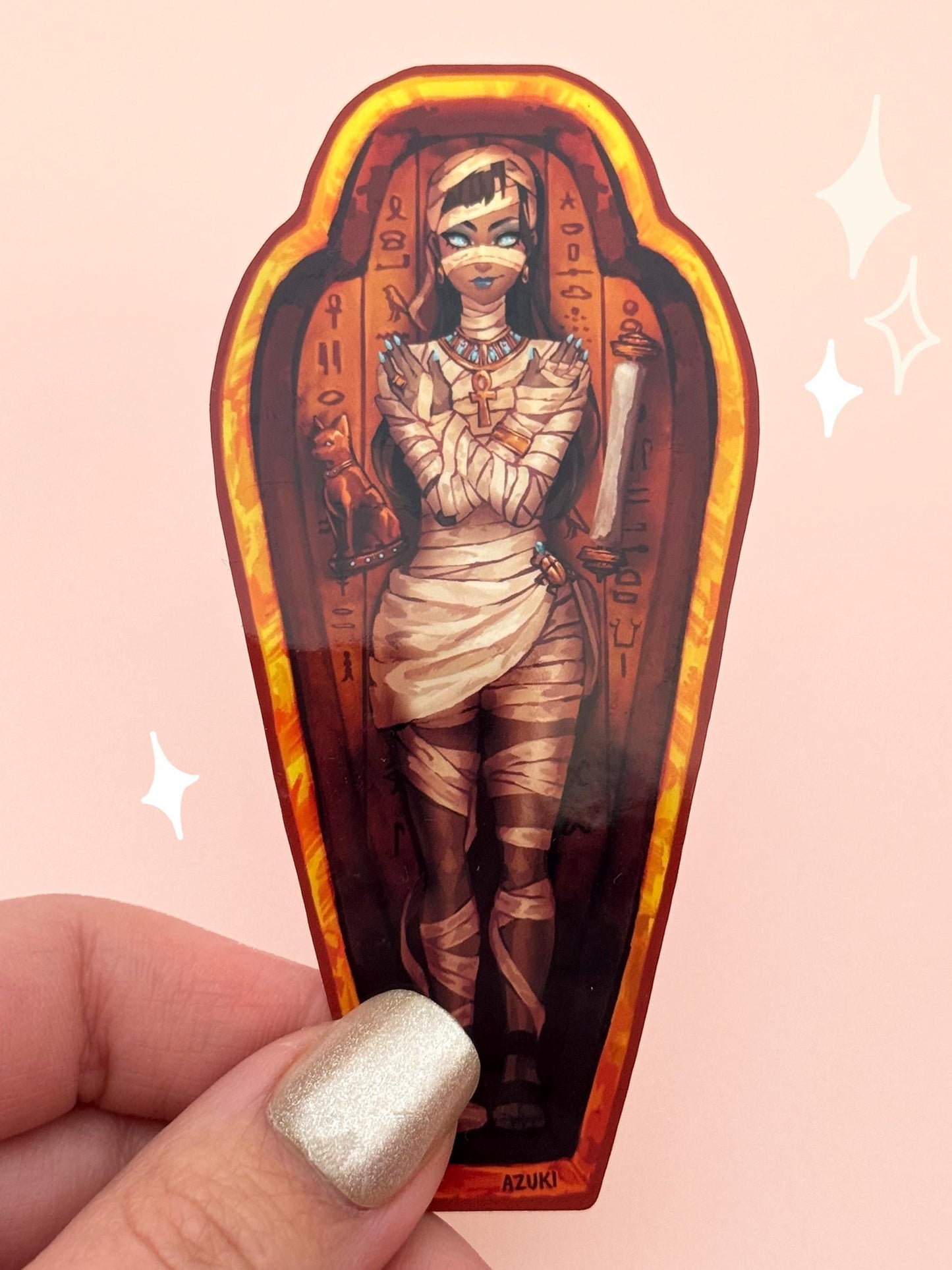 Die-cut vinyl sticker shaped like an Egyptian sarcophagus, with a mummy girl wrapped in bandages, glowing blue eyes, an ankh necklace, arms crossed over her chest, surrounded by golden hieroglyphs, held between fingers against a peach background.
