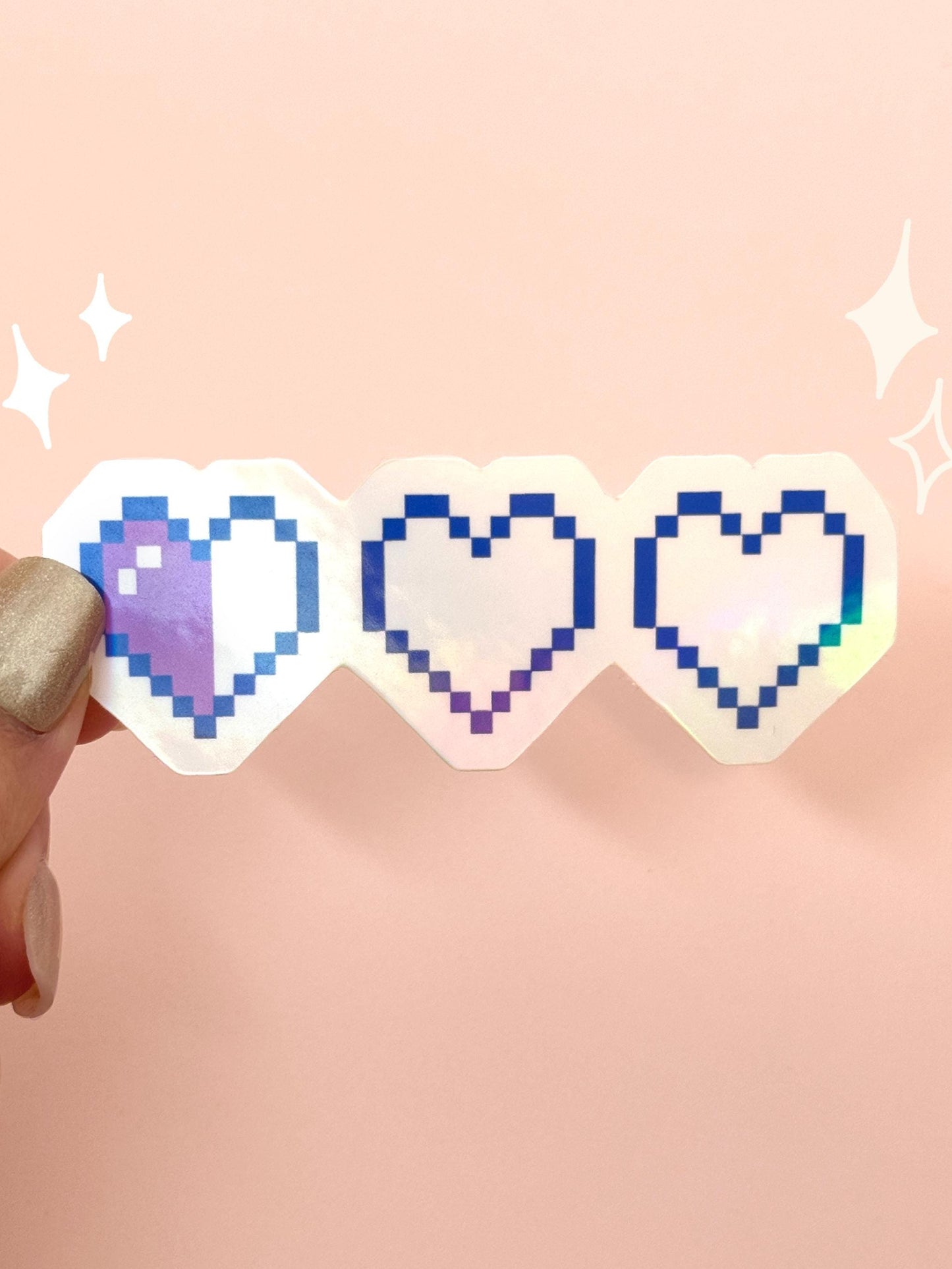 Die-cut holographic vinyl sticker of three pixel hearts in a row depleting from full to empty, like a retro video game health bar, in blue pixel outlines with an iridescent finish, held between fingers against a peach background.
