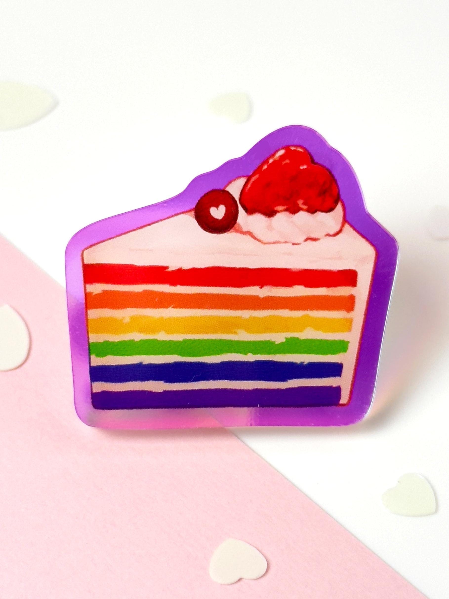 Translucent acrylic pin of a rainbow layer cake slice topped with cream, a strawberry, and a cherry, with an iridescent rainbow border, on a pink and white heart-dotted background.