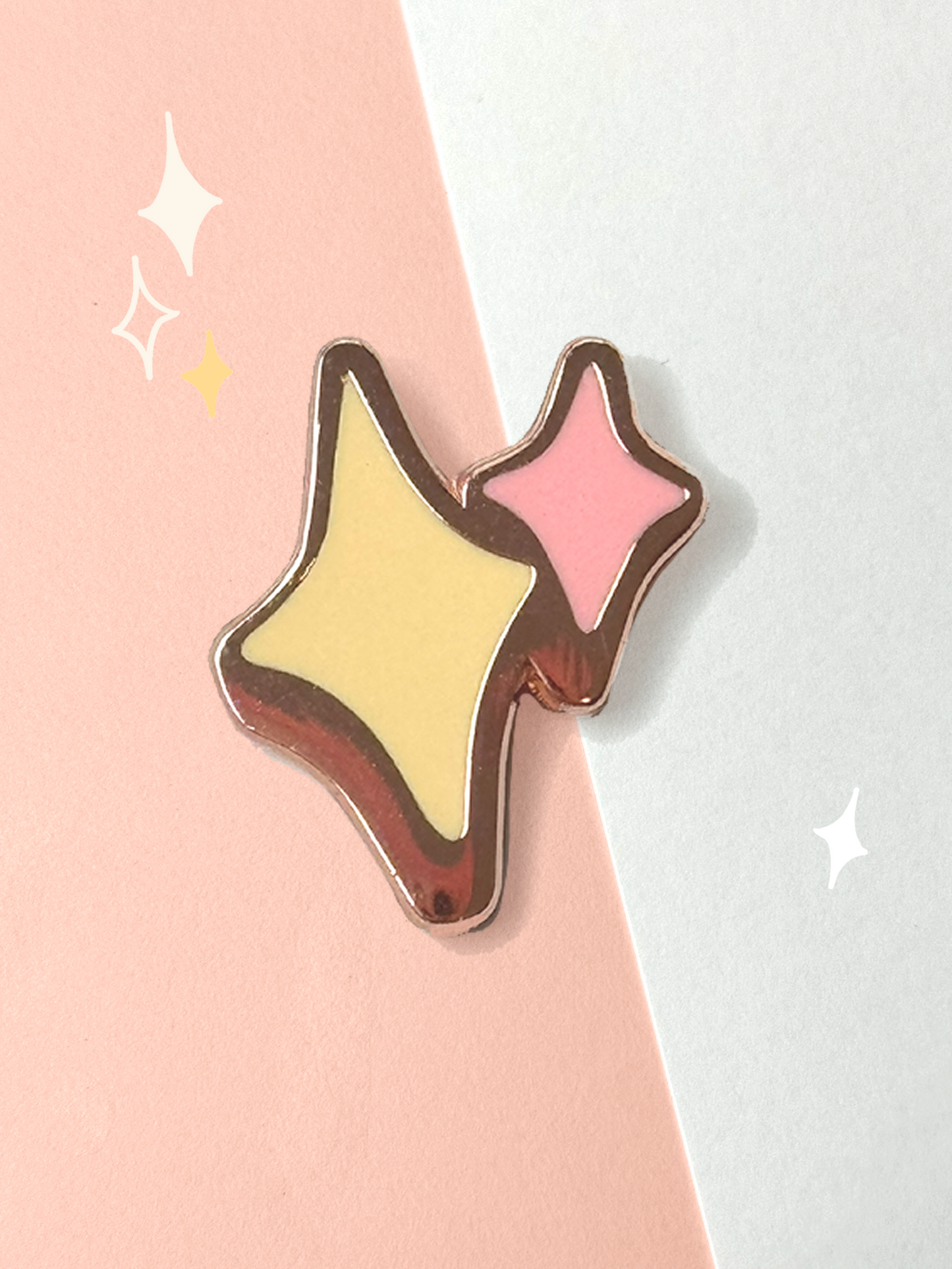 Hard enamel pin of two four-point sparkle stars, one yellow and one pink, outlined in rose-gold metal lines on a pink and white background.