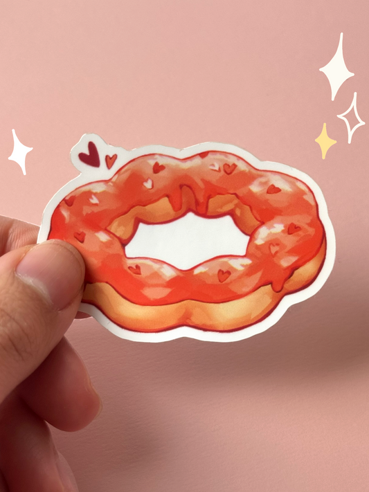 Die-cut soft-touch vinyl sticker of a pon-de-ring style mochi donut with glossy strawberry glaze and tiny heart sprinkles, held between fingers against a pink background.