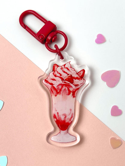 Acrylic keychain shaped like a strawberry parfait in a tall sundae glass — whipped cream, strawberry slices, and strawberry sauce — with a red clasp, on a pink and white background with heart confetti.