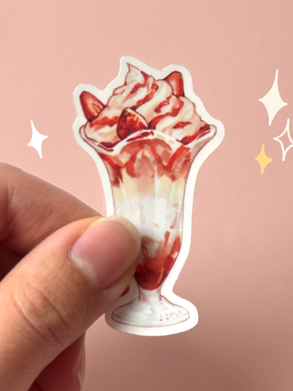 Die-cut soft-touch vinyl sticker of a strawberry parfait in a tall sundae glass — whipped cream, strawberry slices, and strawberry sauce layered to the top, held between fingers against a pink background.