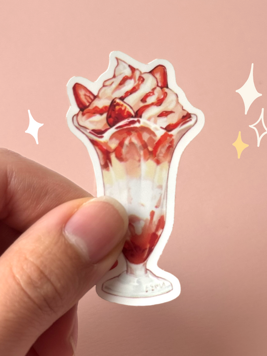 Die-cut soft-touch vinyl sticker of a strawberry parfait in a tall sundae glass — whipped cream, strawberry slices, and strawberry sauce layered to the top, held between fingers against a pink background.