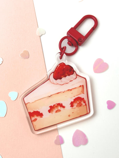 Acrylic keychain shaped like a painterly slice of strawberry shortcake — layered sponge and cream with strawberries inside and one on top, with a red clasp, on a pink and white background with heart confetti.