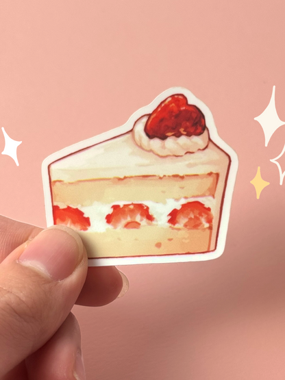 Die-cut soft-touch vinyl sticker of a painterly slice of strawberry shortcake — layered sponge and cream with whole strawberries inside and a strawberry on top, held between fingers against a peach background.