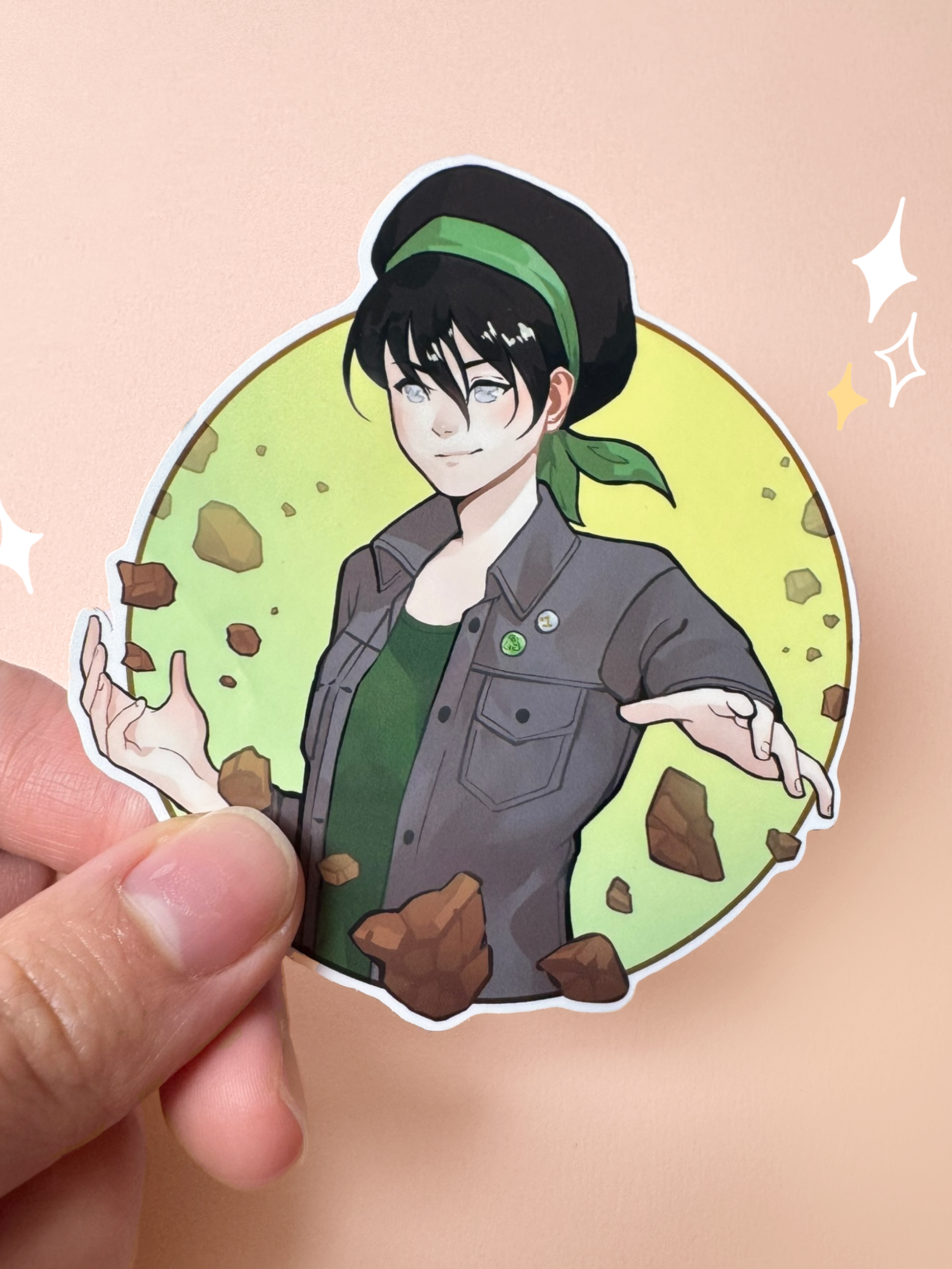 Die-cut vinyl sticker of Toph from Avatar: The Last Airbender in a modern outfit — black hair with her iconic green headband, grey denim jacket over a green tank top, milky pale eyes, with earthbending rocks floating around her in a circular green frame, held between fingers against a peach background.