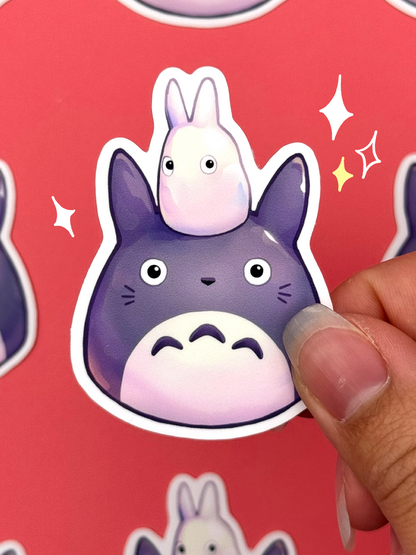Die-cut vinyl sticker of a large grey Totoro with a small white Totoro sitting on its head, both drawn in a round squishy mochi style, held between fingers against a coral background.