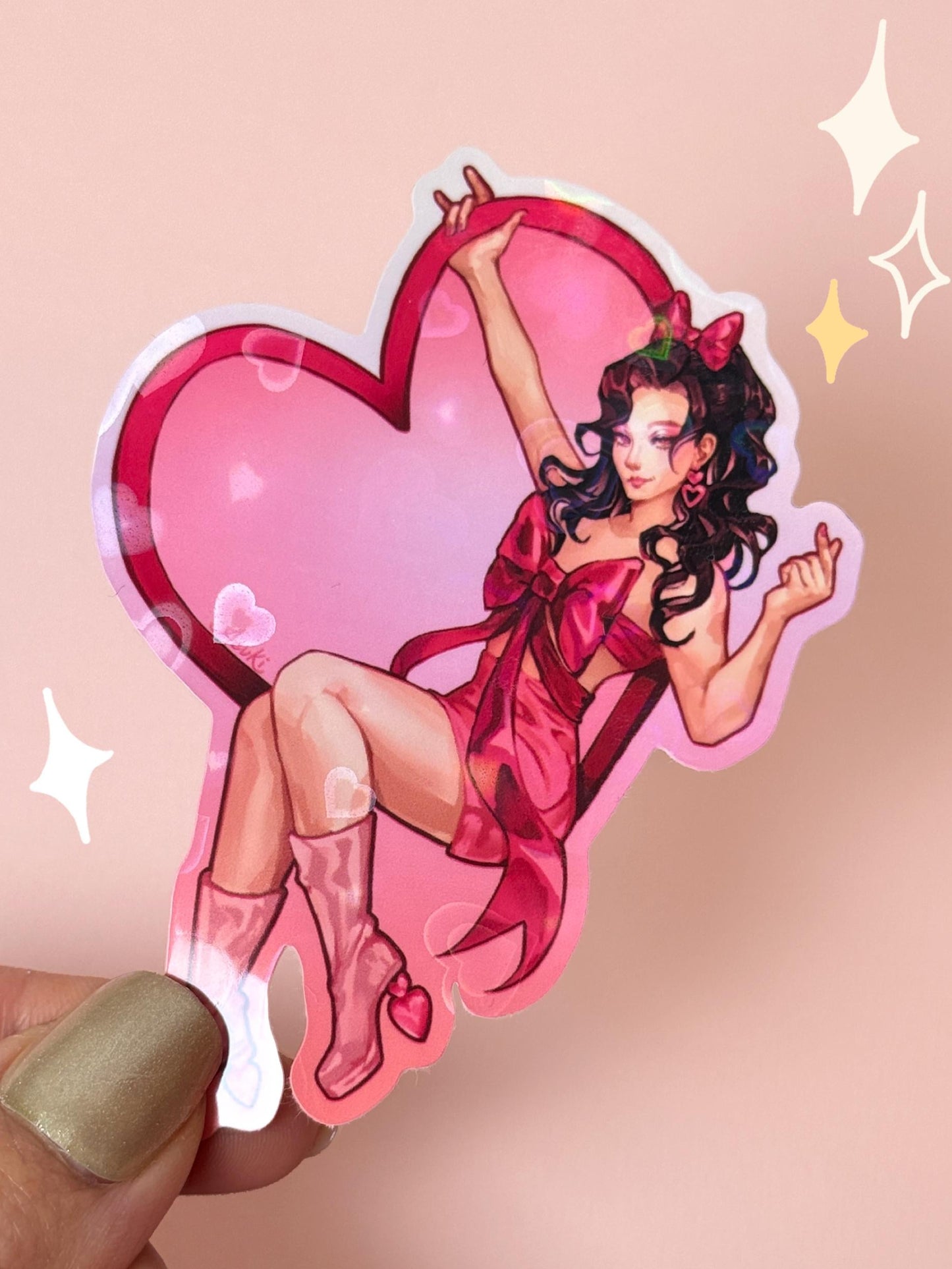 Die-cut holographic vinyl sticker of a pin-up girl with dark curly hair in a red ribbon-wrapped dress and pink boots, posed against a large pink heart with a holographic sparkle finish, held between fingers against a peach background.