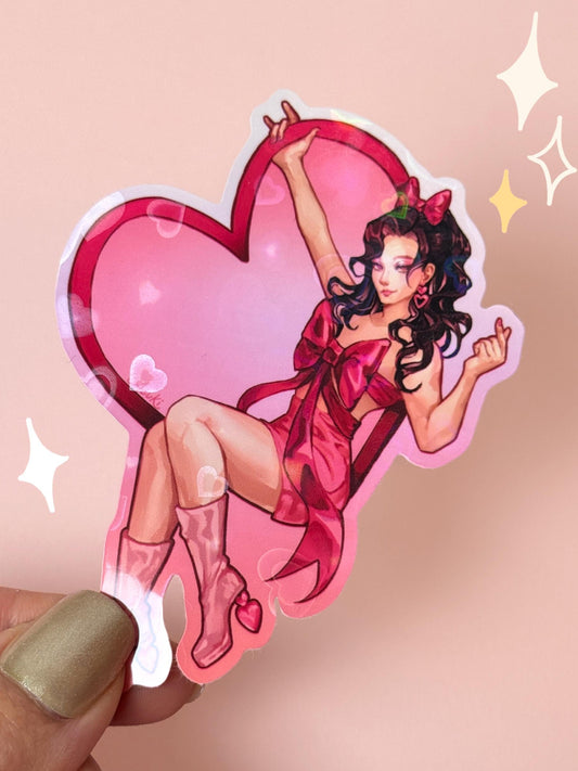 Die-cut holographic vinyl sticker of a pin-up girl with dark curly hair in a red ribbon-wrapped dress and pink boots, posed against a large pink heart with a holographic sparkle finish, held between fingers against a peach background.