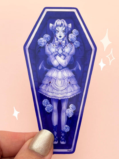Die-cut vinyl sticker shaped like a coffin, with a vampire girl in a bat-inspired gothic lolita dress, pointed ears, bat wings, fangs, and pigtails, surrounded by white roses in a plush-lined coffin, held between fingers against a peach background.