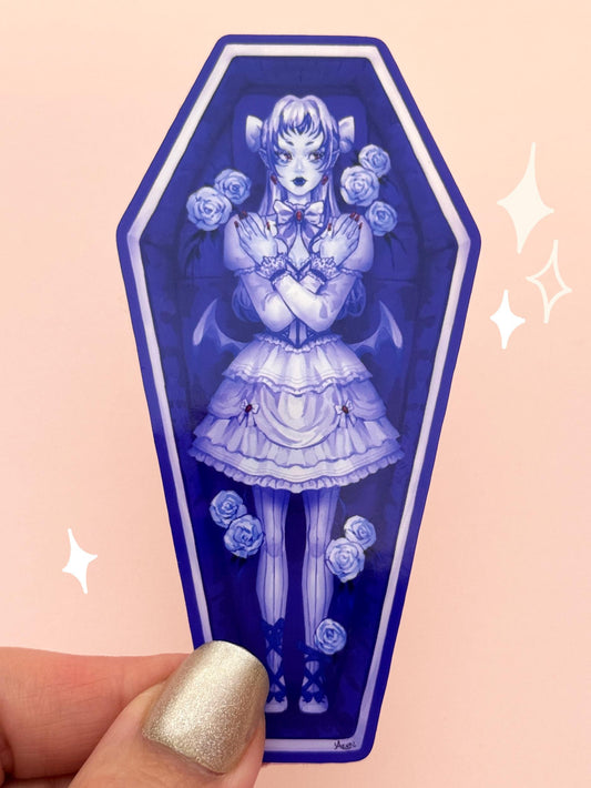 Die-cut vinyl sticker shaped like a coffin, with a vampire girl in a bat-inspired gothic lolita dress, pointed ears, bat wings, fangs, and pigtails, surrounded by white roses in a plush-lined coffin, held between fingers against a peach background.