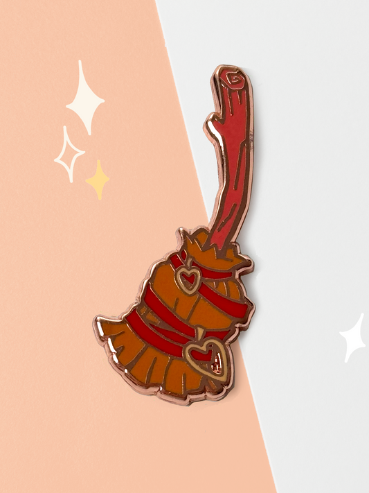 Hard enamel witch broom pin with a red wood handle, an amber-orange bristle skirt bound in red ribbon, and two rose-gold heart charms, outlined in rose-gold metal lines.