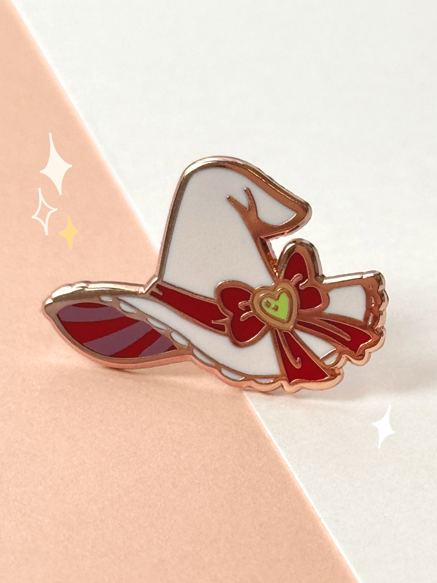 Hard enamel witch hat pin in cream with a red ribbon band, a red bow centred on a green heart charm, a lacy brim, a striped underside, and rose-gold metal lines.