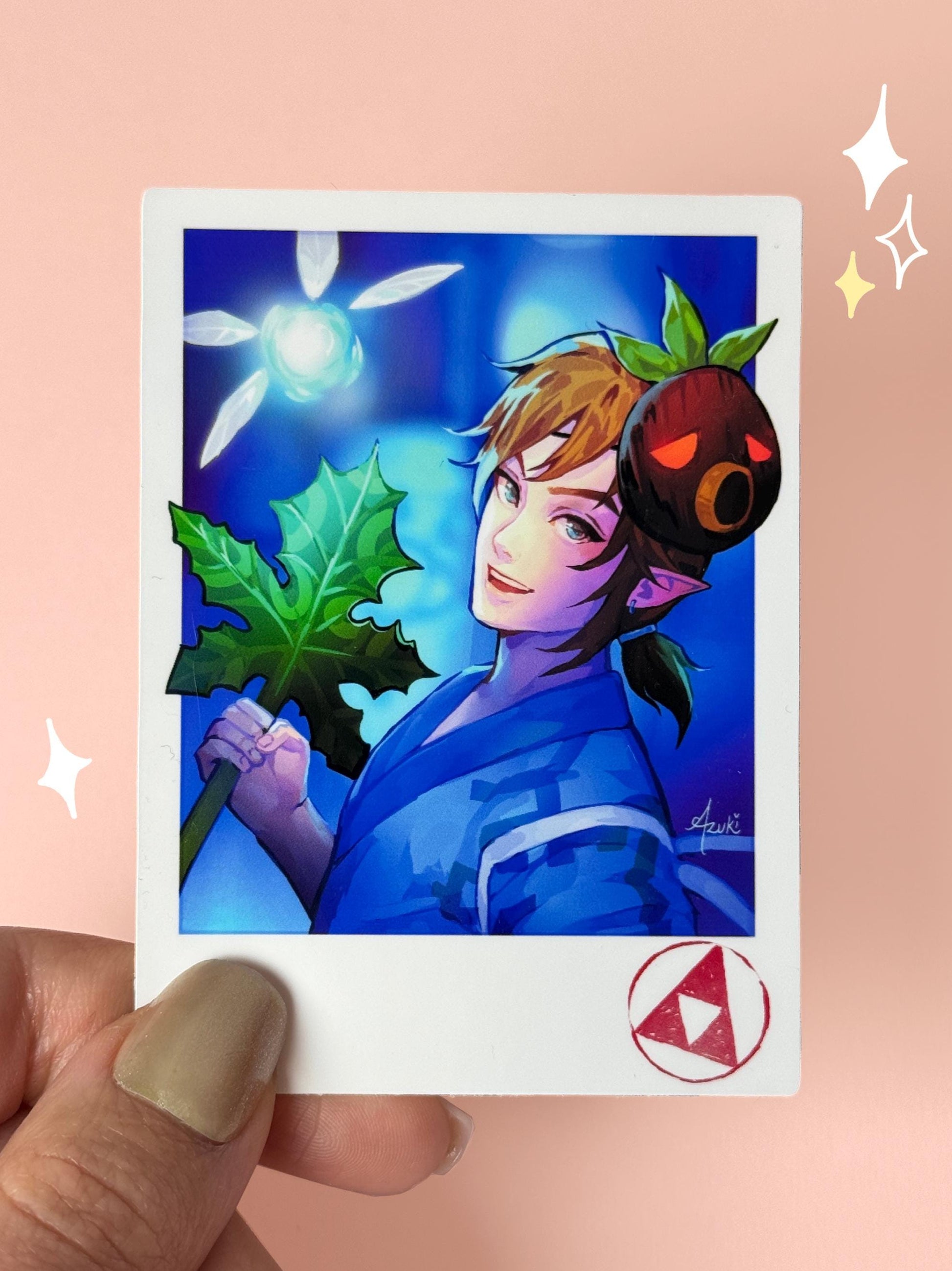 Polaroid-style vinyl sticker of Link from The Legend of Zelda in a blue yukata at a matsuri, holding a Korok leaf, with a Deku mask on his head, a glowing fairy beside him, and a Triforce stamp in the corner, held between fingers against a peach background.