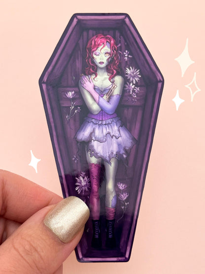 Die-cut vinyl sticker shaped like a wooden coffin, with a zombie girl in a tattered purple Victorian dress, red hair, stitched green-and-purple skin, and mismatched eyes, surrounded by wilting wildflowers, held between fingers against a peach background.