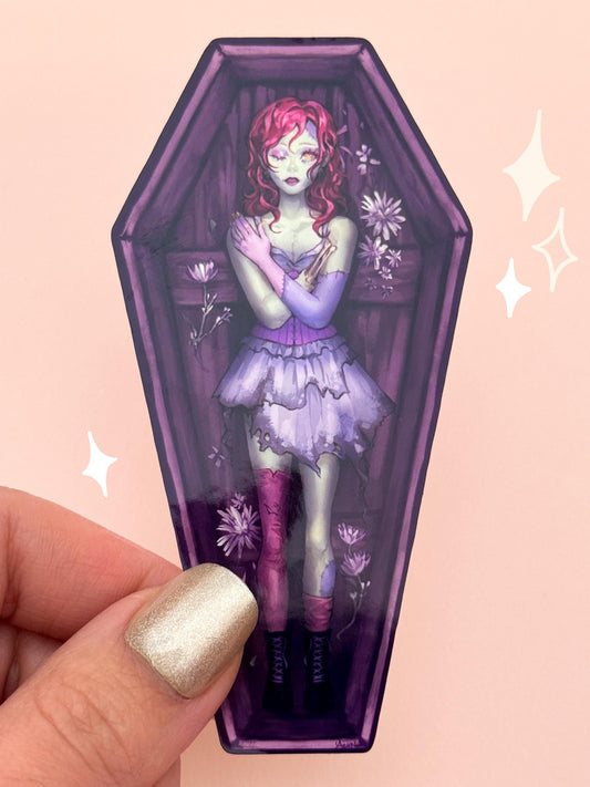 Die-cut vinyl sticker shaped like a wooden coffin, with a zombie girl in a tattered purple Victorian dress, red hair, stitched green-and-purple skin, and mismatched eyes, surrounded by wilting wildflowers, held between fingers against a peach background.
